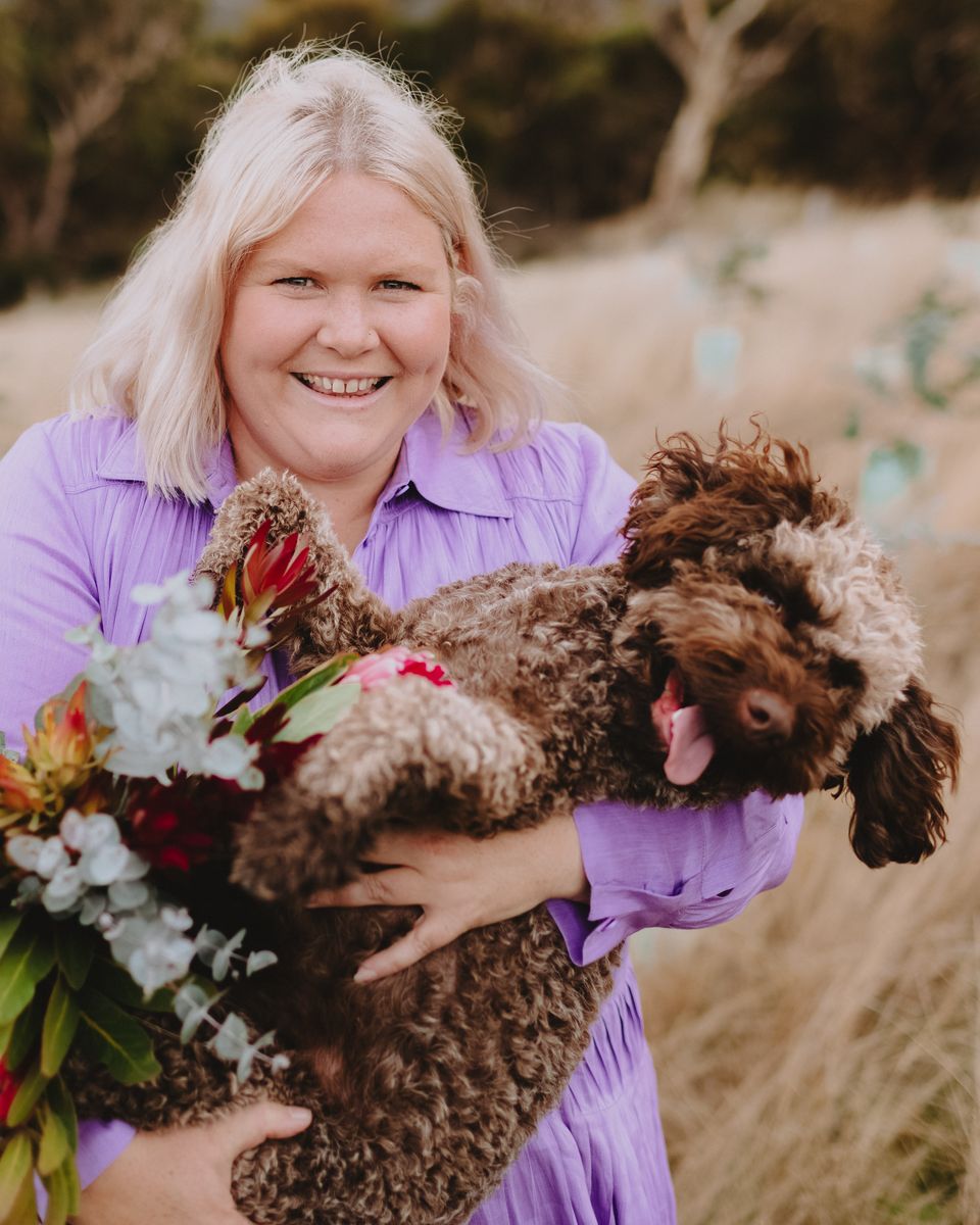 Nikki smiling in a field, holding her dog and a bunch of proteas and eucalyptus