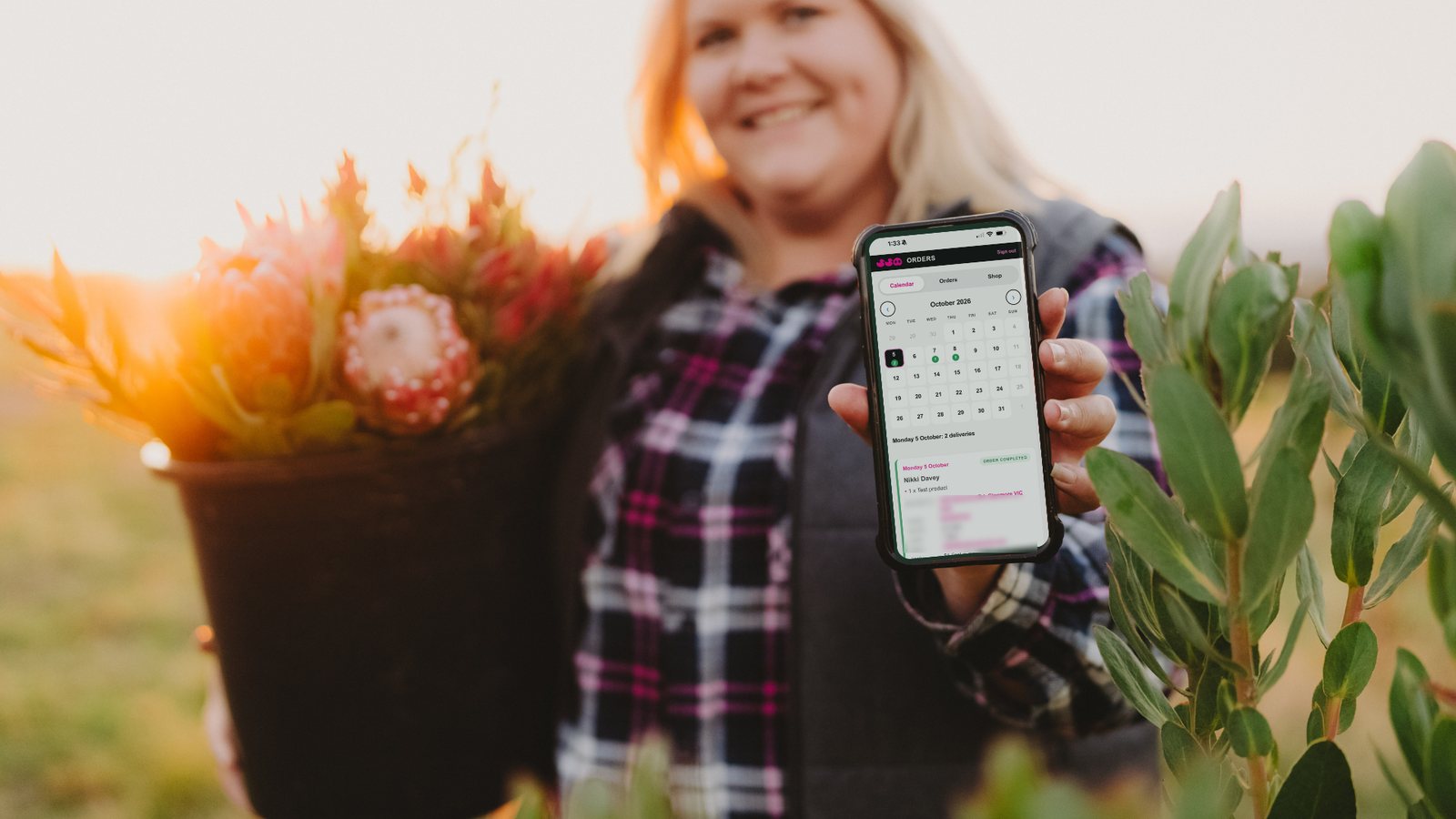 Nikki holding a bucket of proteas in one hand and a phone showing the Duck Duck Pig orders app in the other