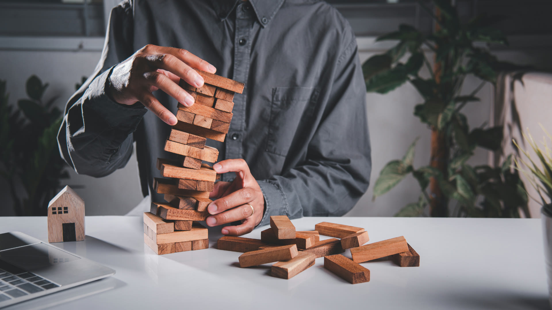 A person steadying a wobbling tower of wooden blocks on a desk, with fallen blocks scattered in front