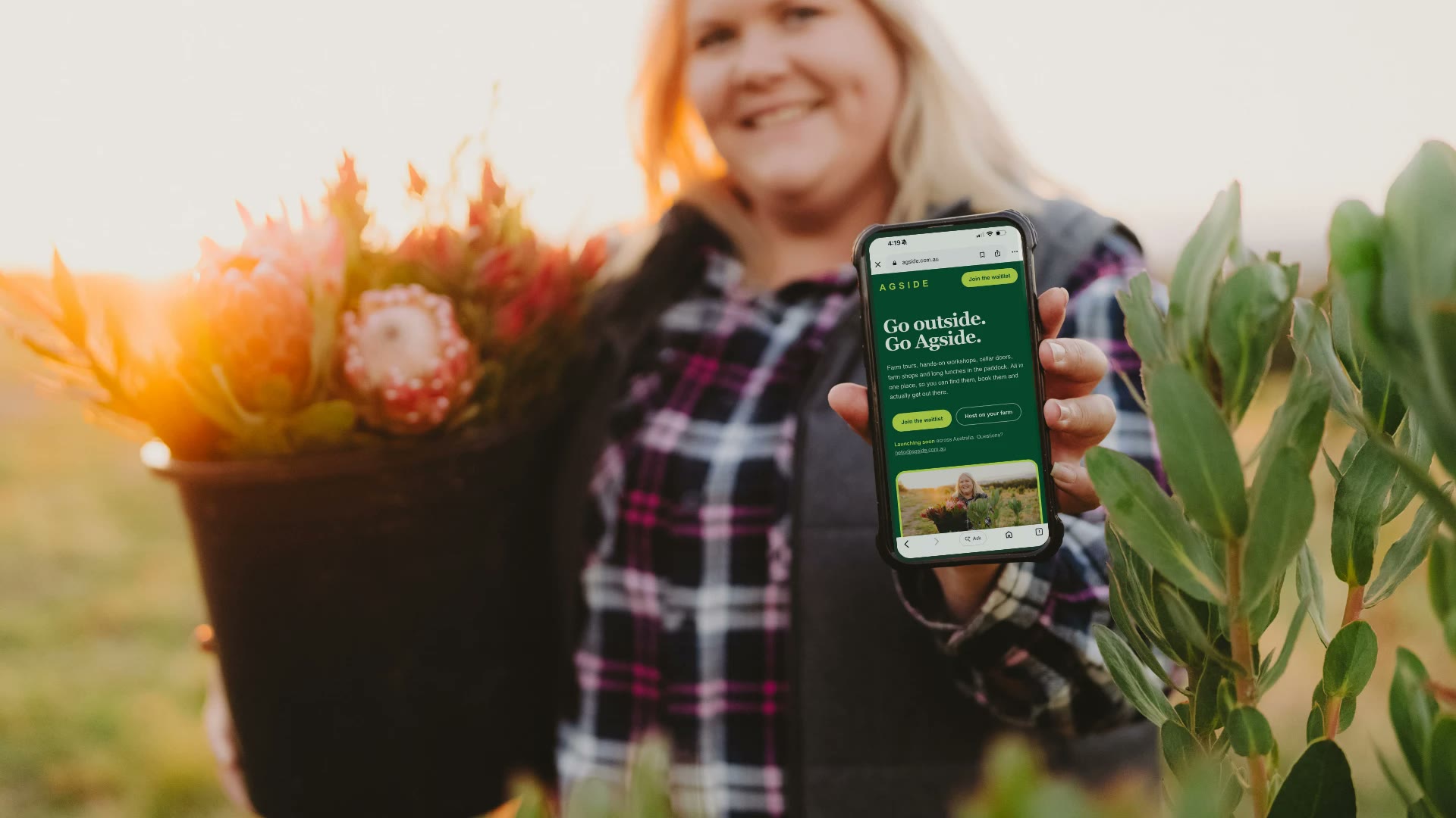 A flower farmer holding a bucket of proteas in one hand and a phone showing the AgSide website in the other