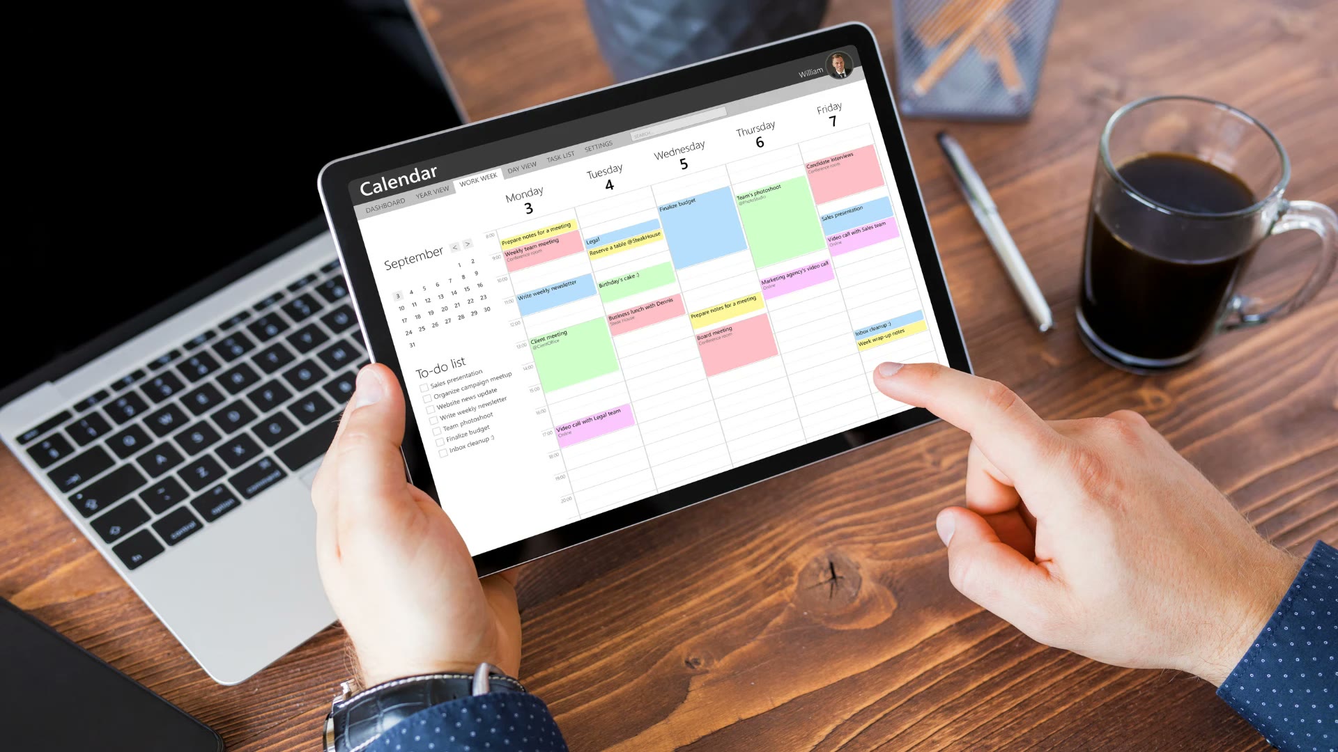 A person holding a tablet showing a colour-coded work-week calendar and to-do list, with a laptop and a coffee on a wooden desk