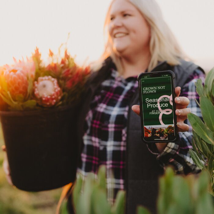 Nikki holding a bucket of proteas and a phone showing the Grown Not Flown app